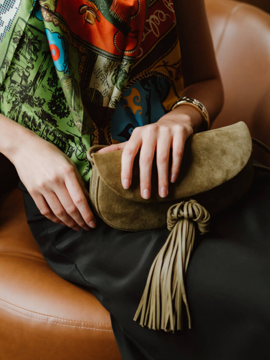 We’re Obsessed With This Rhinestone Cowboy Clutch (and 5 Other Nashville Fall Styles) - 5 A person sitting on a brown leather chair, wearing a colorful scarf and black skirt, holds a green suede clutch bag with a large tassel—an accessory found in trendy Nashville boutiques.