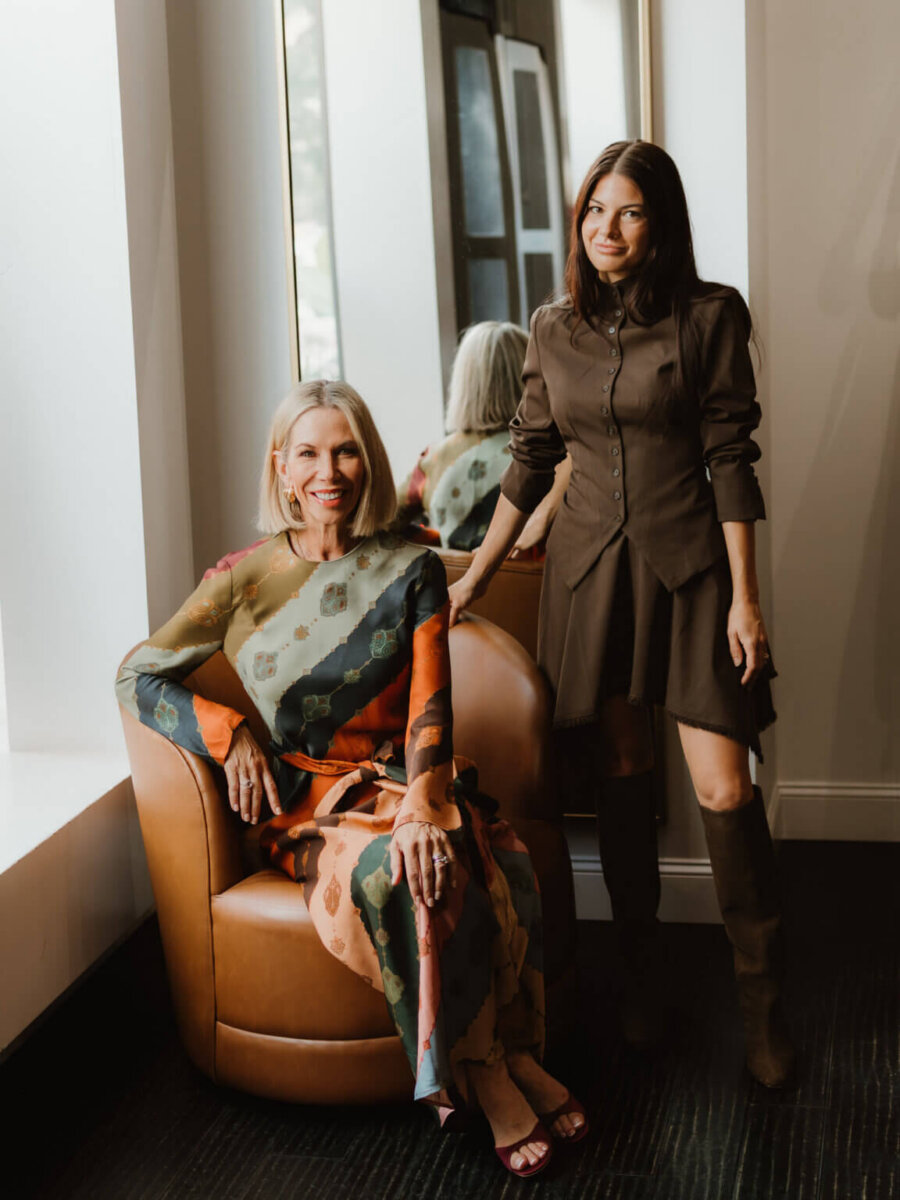 We’re Obsessed With This Rhinestone Cowboy Clutch (and 5 Other Nashville Fall Styles) - 6 Two women pose indoors by a window; one is seated in a brown chair wearing a colorful dress from one of the trendiest Nashville boutiques, and the other stands beside her in a brown dress and knee-high boots.