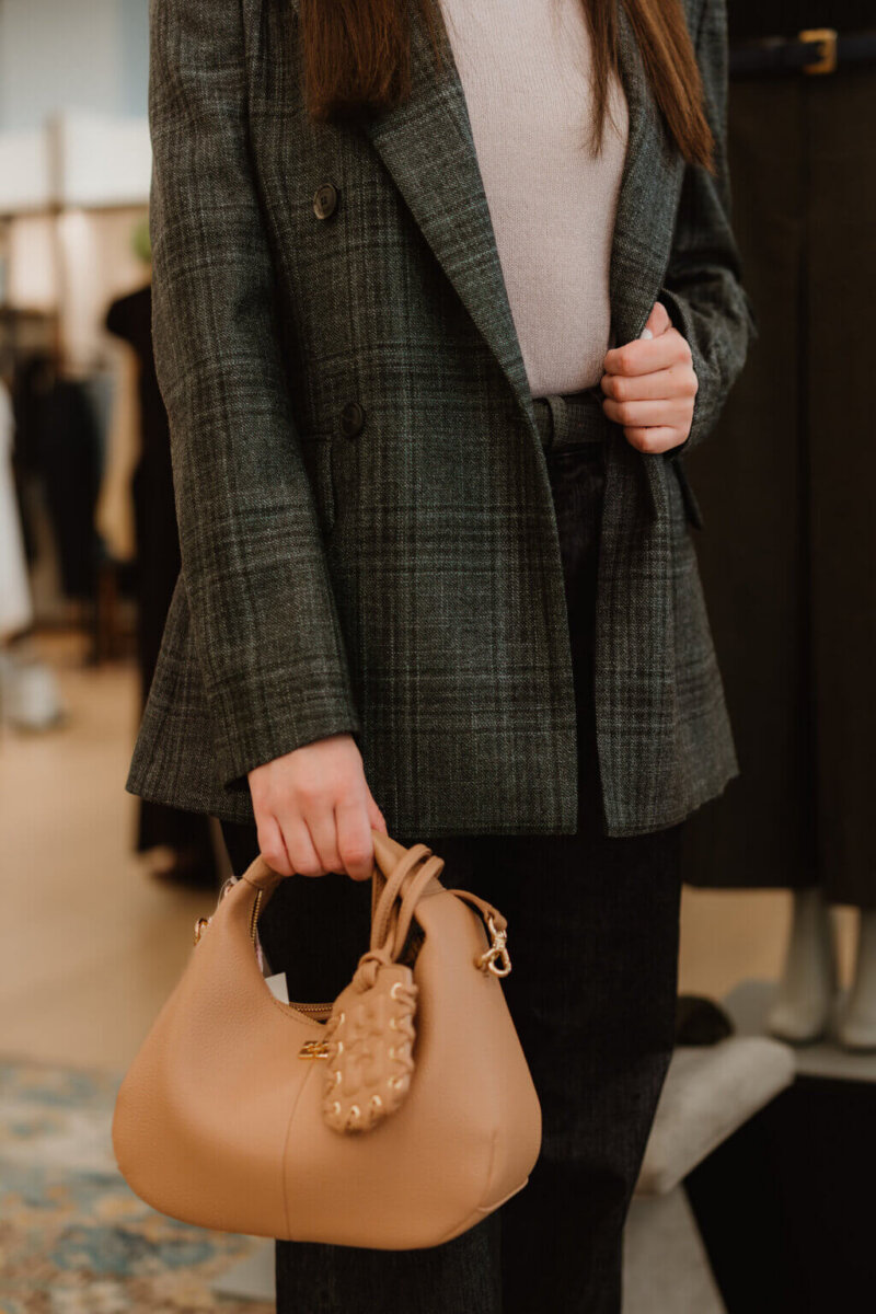 We’re Obsessed With This Rhinestone Cowboy Clutch (and 5 Other Nashville Fall Styles) - 8 Person wearing a plaid blazer and light sweater holds a beige handbag with decorative details, standing indoors near clothing displays often found in stylish Nashville boutiques.