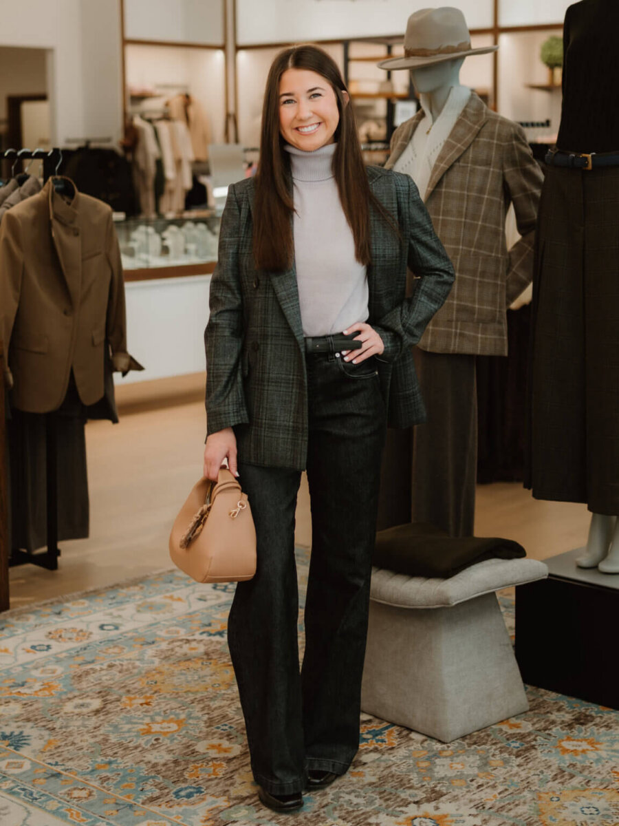 We’re Obsessed With This Rhinestone Cowboy Clutch (and 5 Other Nashville Fall Styles) - 7 Woman stands indoors wearing a plaid blazer, gray turtleneck, dark pants, and holding a tan handbag. Mannequins in business attire and clothing racks are visible in the background.