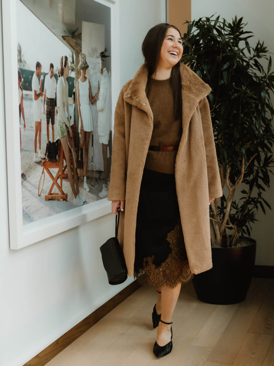We’re Obsessed With This Rhinestone Cowboy Clutch (and 5 Other Nashville Fall Styles) - 9 A woman in a brown coat, brown sweater, and black skirt stands indoors, smiling, holding a black handbag. There is a large framed photo and a potted plant behind her.