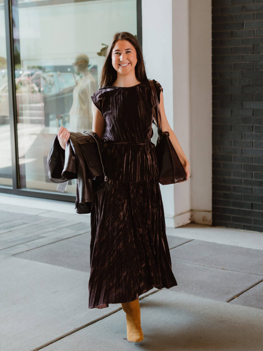 We’re Obsessed With This Rhinestone Cowboy Clutch (and 5 Other Nashville Fall Styles) - 10 Woman in a dark pleated dress and tan boots walks outside, holding a jacket in each hand, smiling near a window and brick wall.