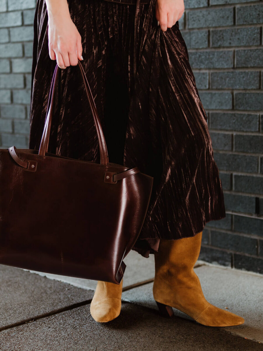 We’re Obsessed With This Rhinestone Cowboy Clutch (and 5 Other Nashville Fall Styles) - 11 Person wearing a dark pleated skirt and tan suede boots, holding a large dark brown leather tote bag from one of the chic Nashville boutiques, standing by a black brick wall.