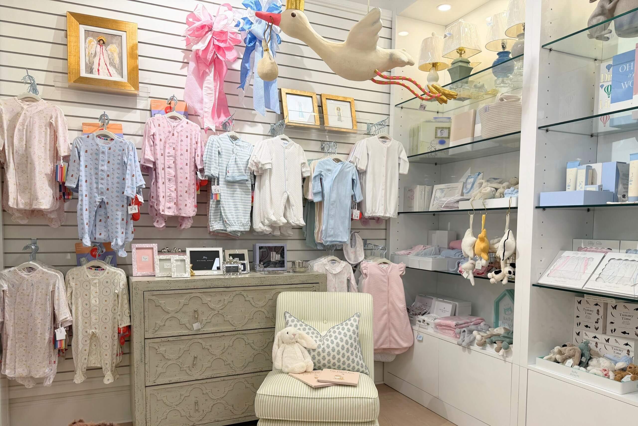 A nursery store display with baby clothes on a slat wall, a dresser, a striped chair with a plush bunny, and shelves of unique home decor gifts.