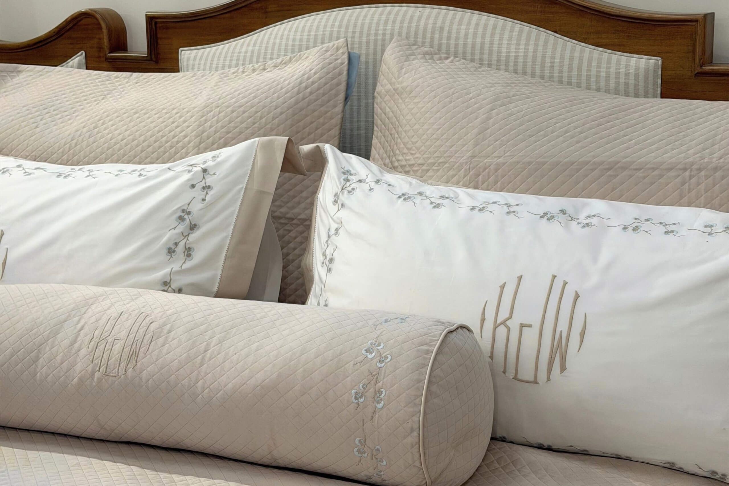 A made bed featuring home decor with quilted beige pillows and white pillowcases embroidered with floral patterns and monograms.
