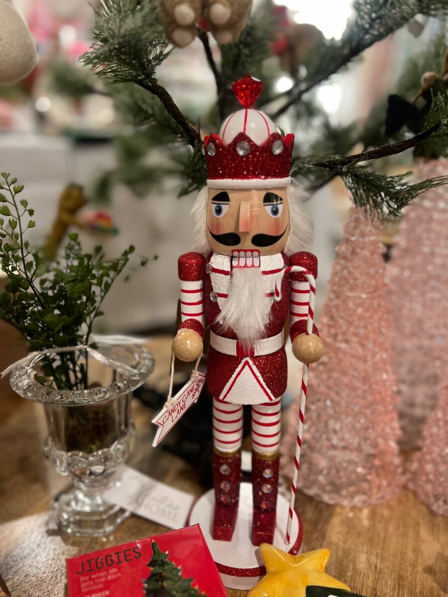 Nashville Steals, Deals & Announcements: October 2026 - 8 A red and white nutcracker figurine stands on a wooden surface beside pink trees, greenery in a glass vase, and a red booklet with “JIGGIES” on the cover.
