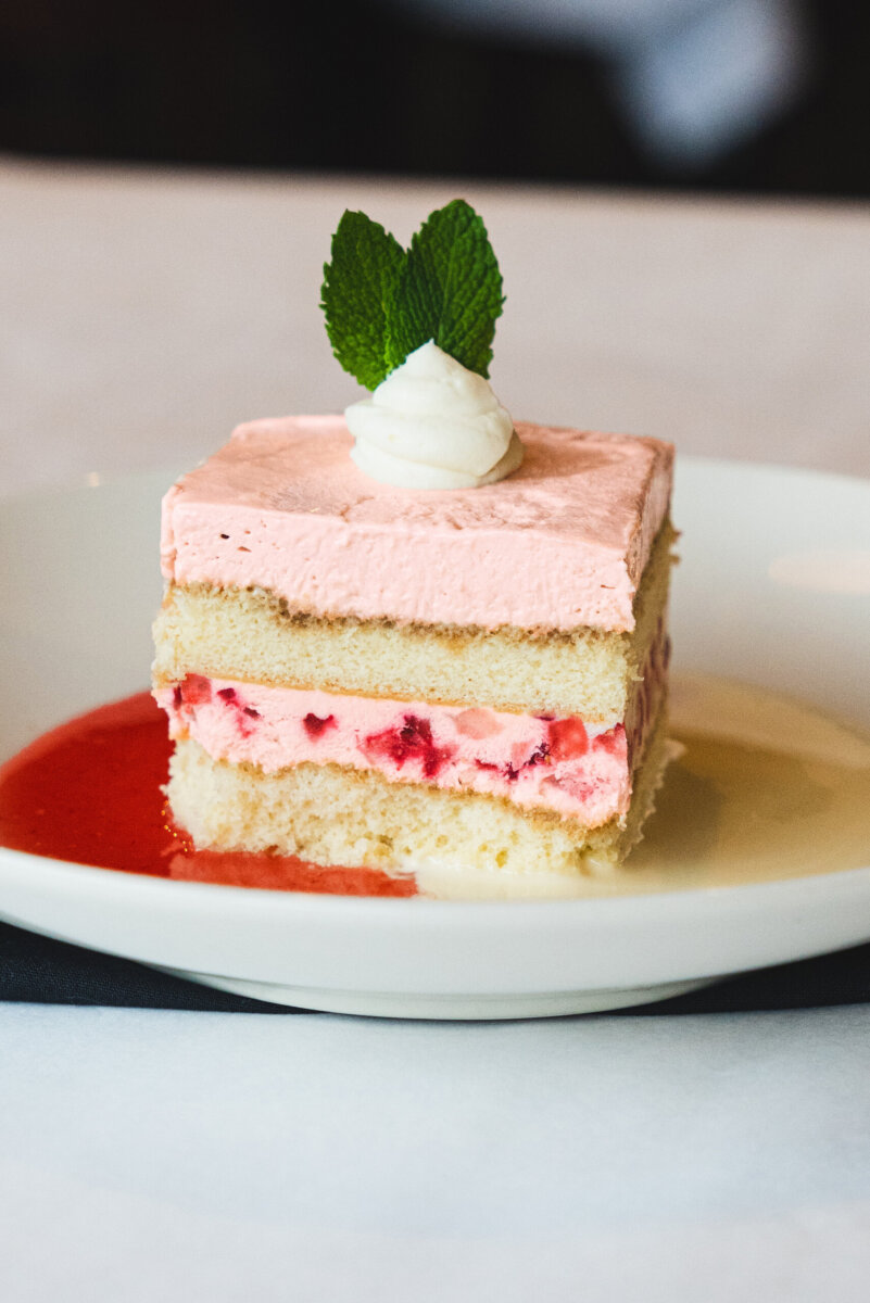 Nashville Steals, Deals & Announcements: October 2026 - 7 A square slice of layered strawberry cake with pink frosting and cream, topped with mint leaves, sits on a white plate with strawberry sauce.