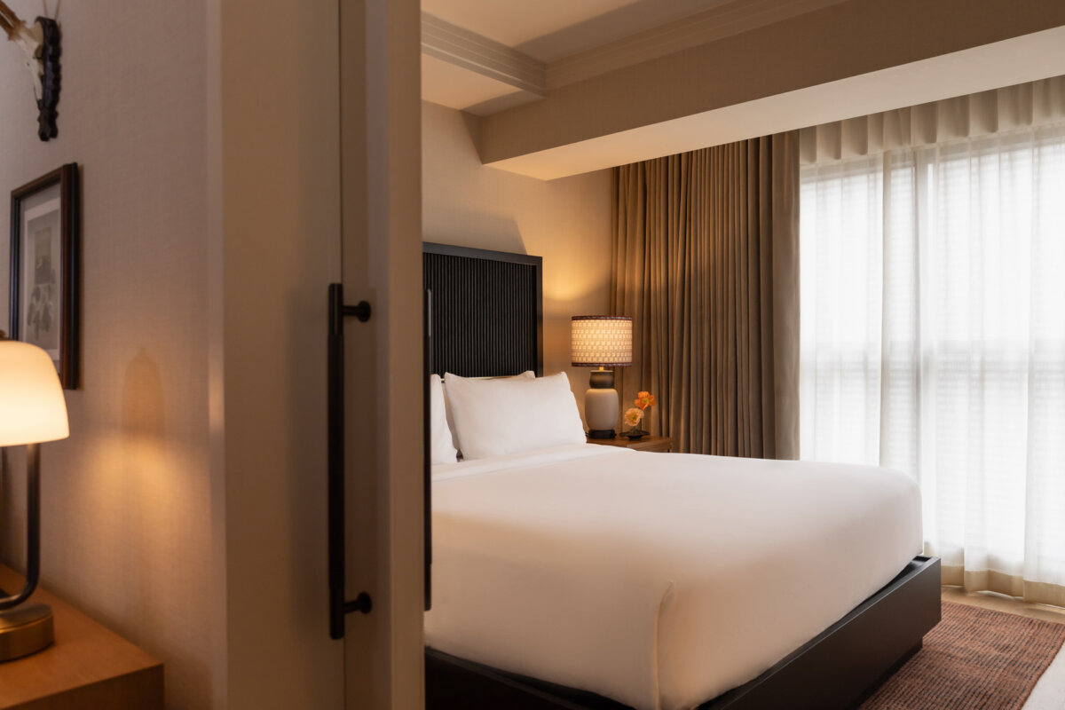 Nashville Steals, Deals & Announcements: October 2026 - 9 A neatly made bed in a hotel room with a bedside lamp, curtains, and soft natural light coming through the window.