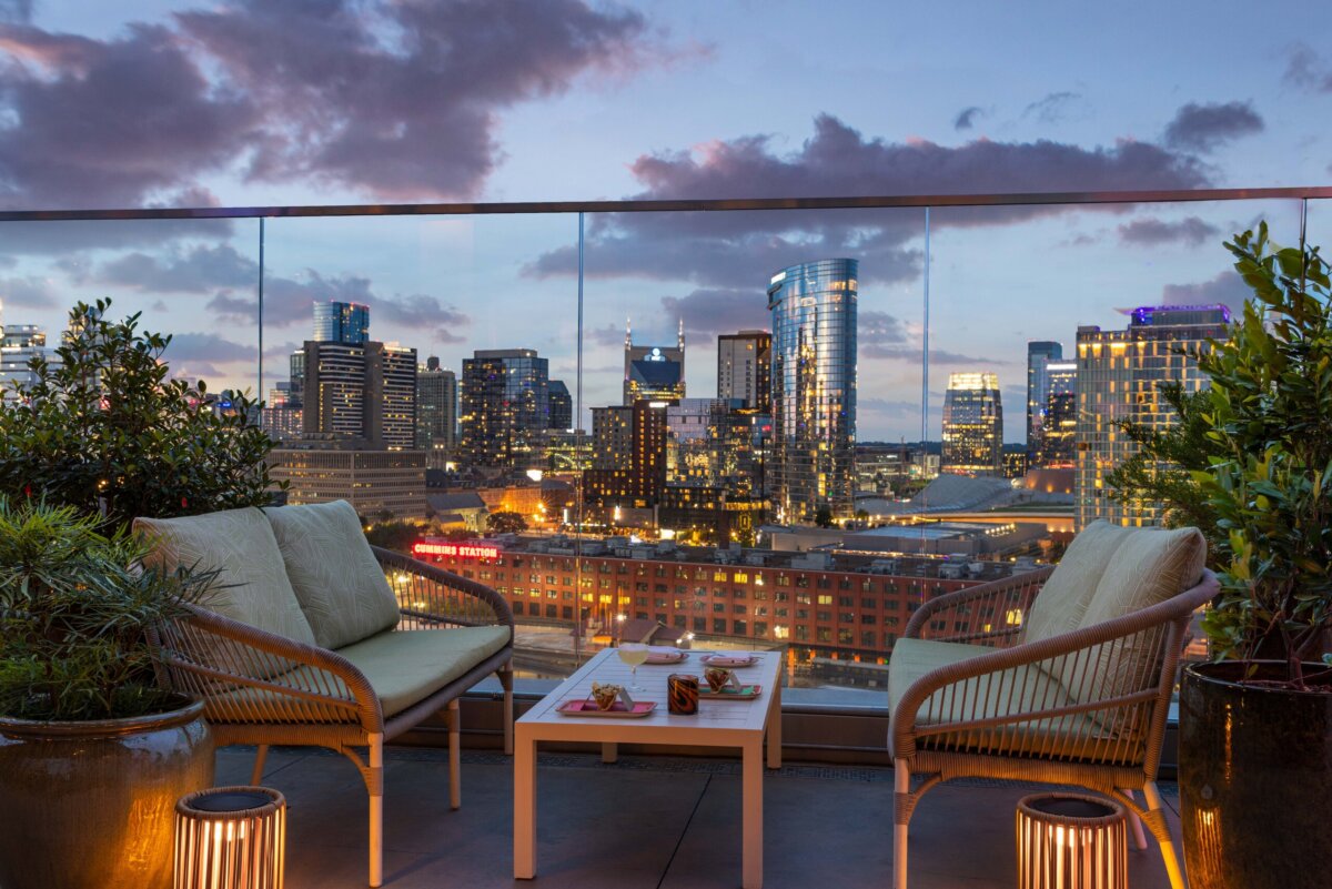 Nashville Steals, Deals & Announcements: October 2026 - 5 Two chairs and a table with drinks and snacks sit on a rooftop patio overlooking a city skyline at dusk, with glass railing and plants around.