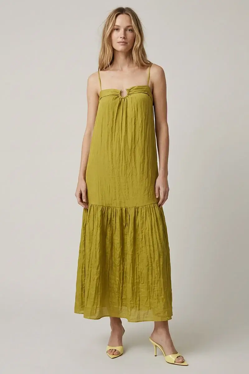 Woman standing in a studio setting wearing a long, yellow-green sleeveless dress with thin straps and ruffled hem, paired with yellow heeled sandals.