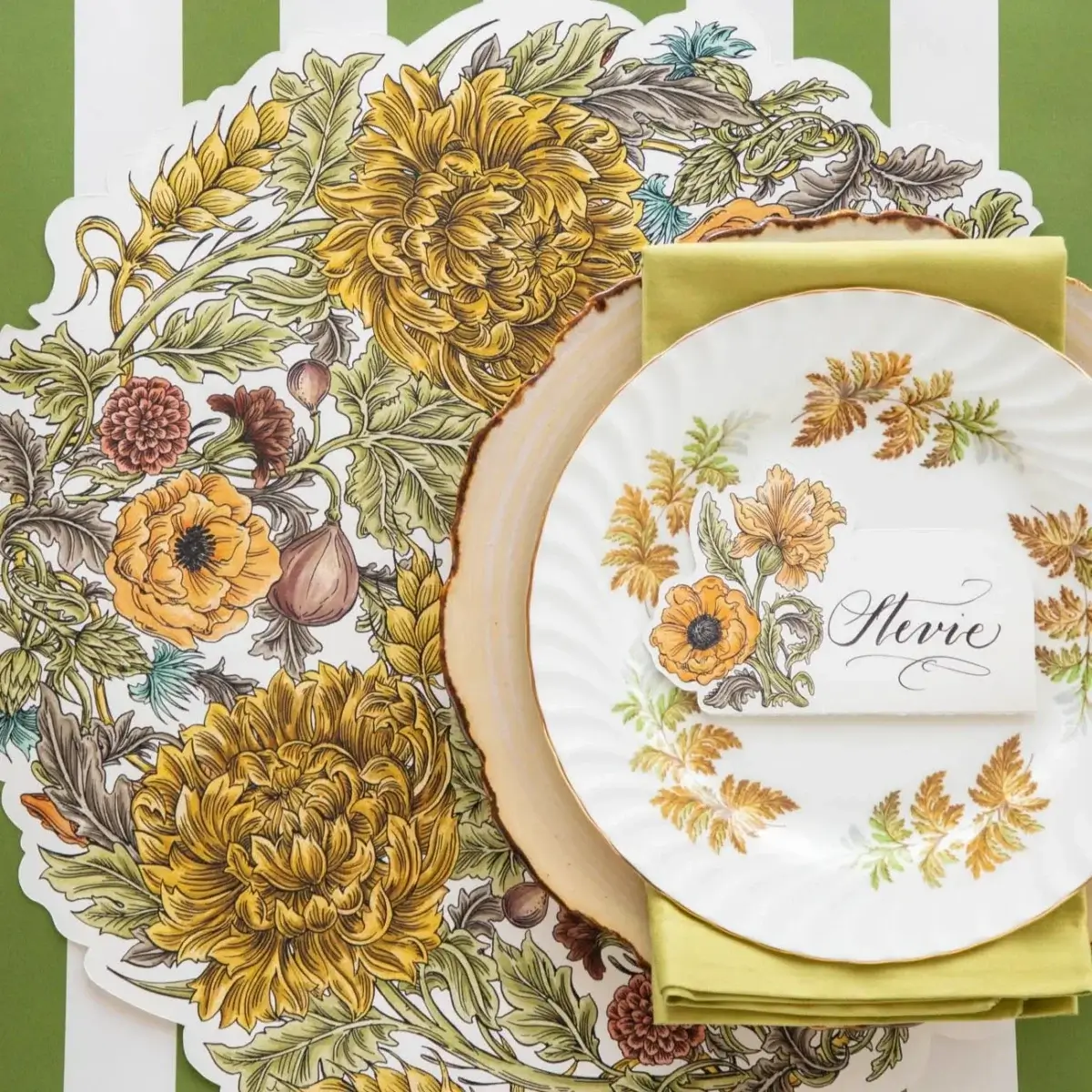 A floral-themed place setting with a decorative placemat, a plate featuring yellow flowers, a green napkin, and a name card reading “Olivia” on a green and white striped surface—perfect inspiration for your next September gathering in Nashville.