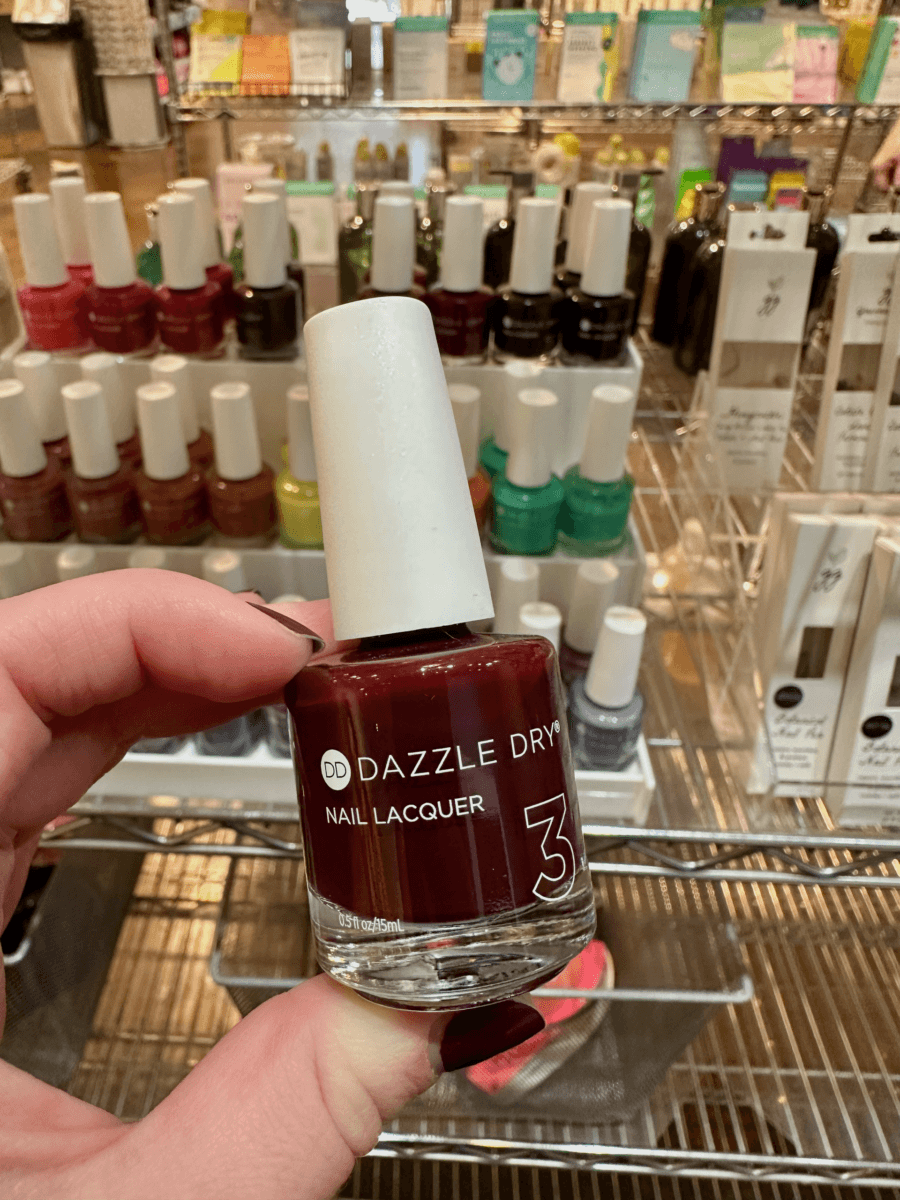 A hand holds a bottle of Dazzle Dry nail lacquer in a store, with shelves of various nail polish bottles in the background.