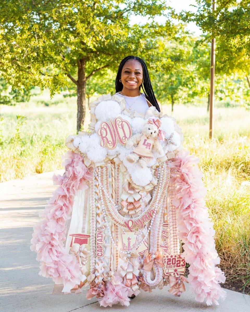 The Bigger the Better! How Texas Homecoming Mums Got So Huge - 2 Smiling girl stands outdoors wearing a large, elaborate pink and white mum adorned with ribbons, flowers, bears, and “2023” decorations—a perfect example of the Texas homecoming mums that celebrate this beloved Texas tradition. Trees and sunlight fill the background.