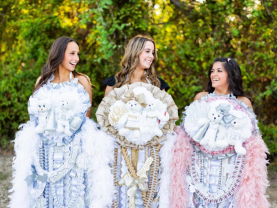 Three young women stand outdoors, each wearing large decorative Texas Homecoming Mums adorned with stuffed animals, ribbons, and trinkets—a colorful display of the cherished Texas tradition. Smiling and looking at each other, they embody the spirit of homecoming.