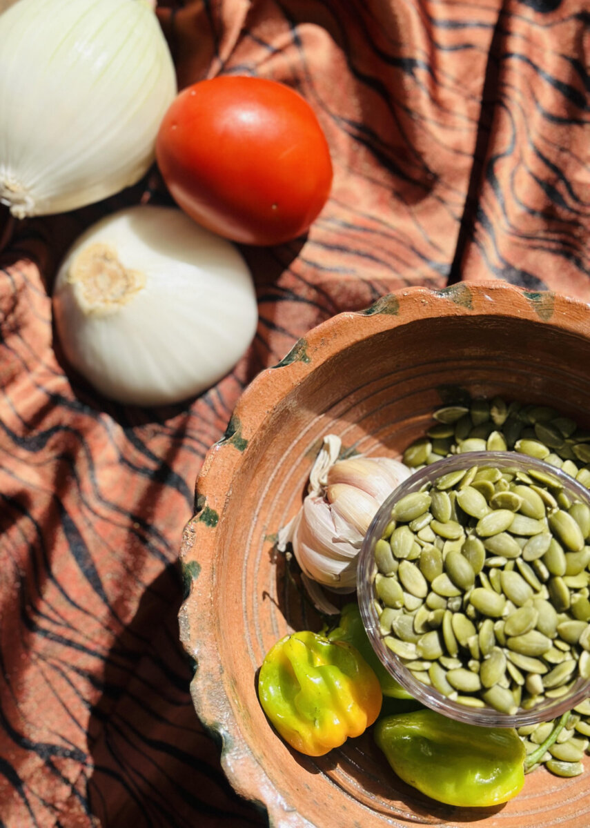 You've GOT to Try This Smoky, Creamy Mexican Dip - 2 A bowl with pumpkin seeds, garlic, and peppers sits on patterned fabric beside whole onions and a tomato—perfect ingredients for crafting a flavorful Mexican dip that’s sure to be a crowd-pleaser on game day.