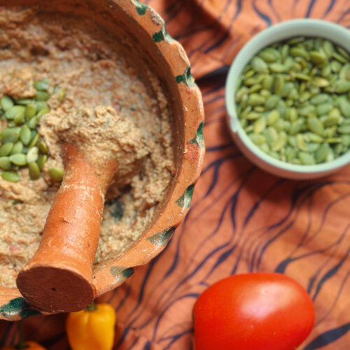 You've GOT to Try This Smoky, Creamy Mexican Dip - 4 A clay mortar with a pestle contains a grainy Mexican dip topped with pumpkin seeds. Nearby are a bowl of pumpkin seeds, two yellow peppers, and a tomato on a patterned cloth—perfect for adding an unexpected dip to your game day spread.