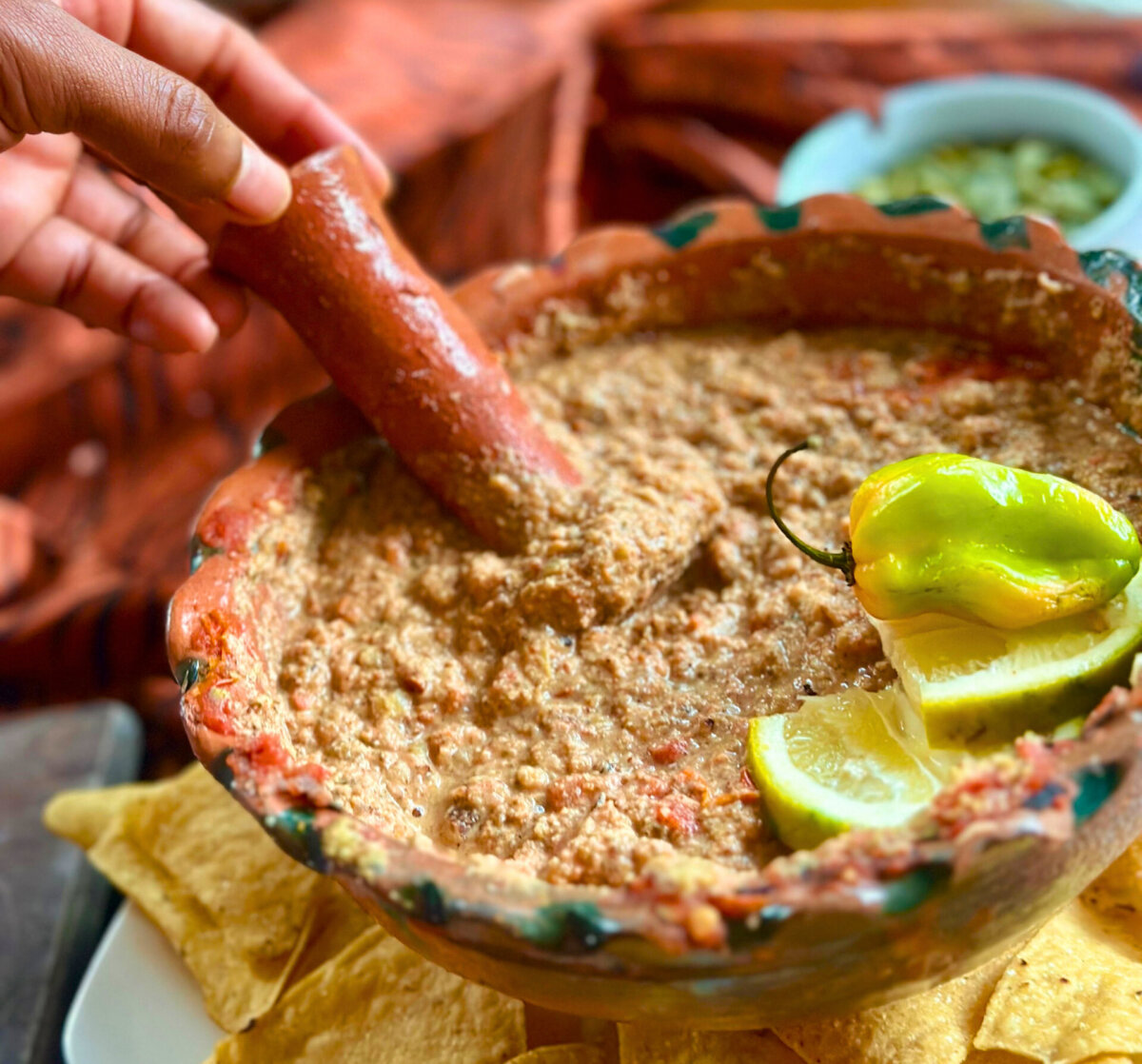 You've GOT to Try This Smoky, Creamy Mexican Dip - 3 A hand dips a utensil into a bowl of chunky Mexican dip, garnished with lemon slices and a pepper, with tortilla chips arranged around the bowl—perfect for Game Day snacking.