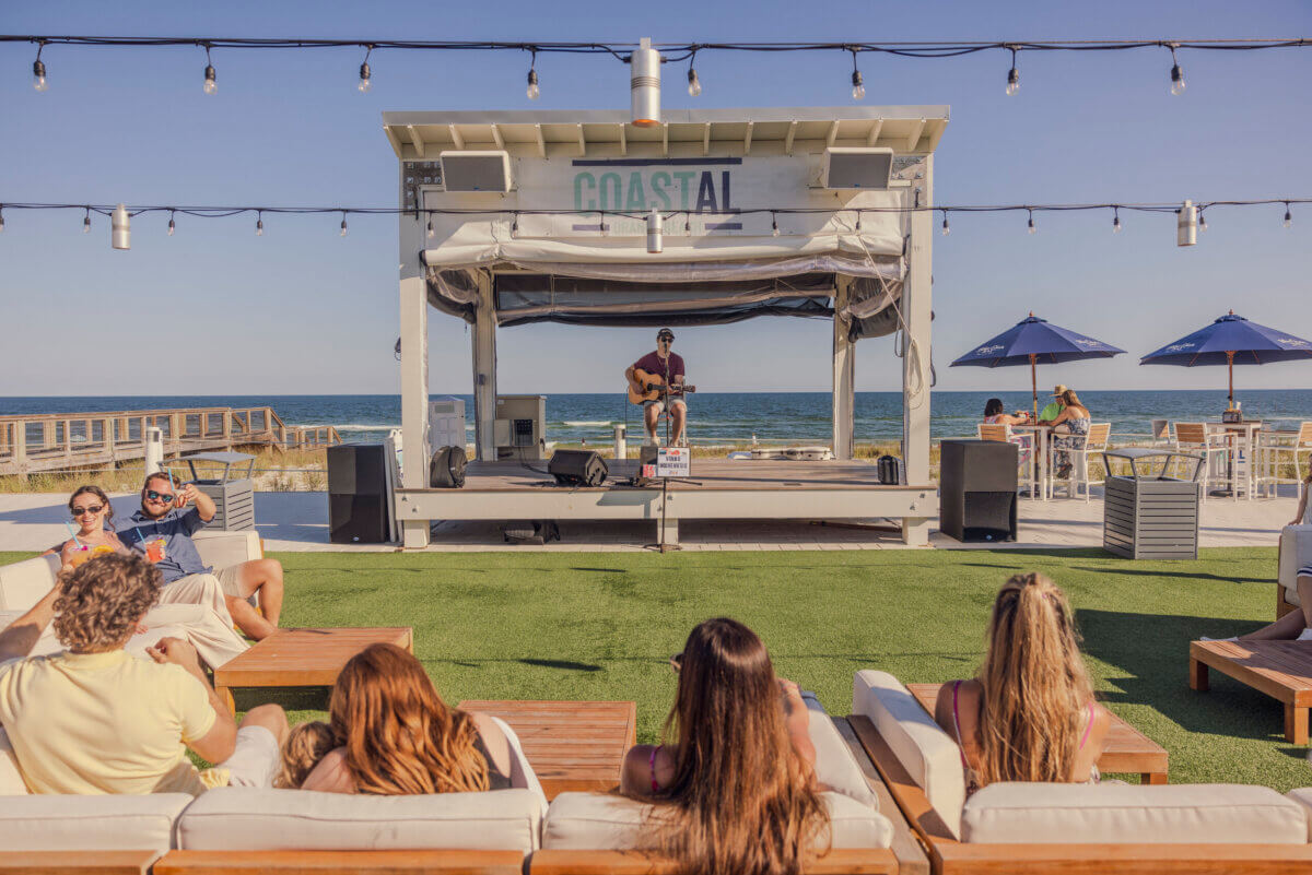 A musician plays guitar on an outdoor stage labeled “Coastal” by the ocean as people relax on couches and chairs under string lights, capturing the laid-back charm of Alabama getaways.