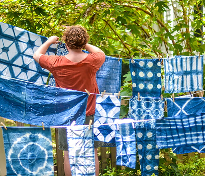 A person hangs various blue and white patterned fabric pieces on clotheslines outdoors among green foliage, evoking the relaxed charm often found on Alabama getaways.