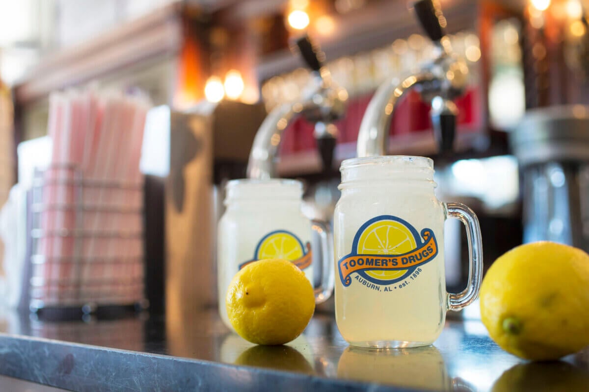 Two mason jars of lemonade with the “Toomer’s Drugs” logo sit on a counter next to fresh lemons and a container of straws, with drink taps blurred in the background—a quintessential taste of Alabama getaways.