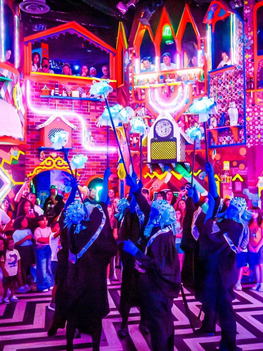 7 Southern Art Museums & Installations Worth Planning a Trip Around - 7 Performers in dark costumes and blue masks raise staffs with glowing objects, surrounded by a crowd in a brightly lit, colorful room with geometric and whimsical decorations at a Meow Wolf Art Installation.