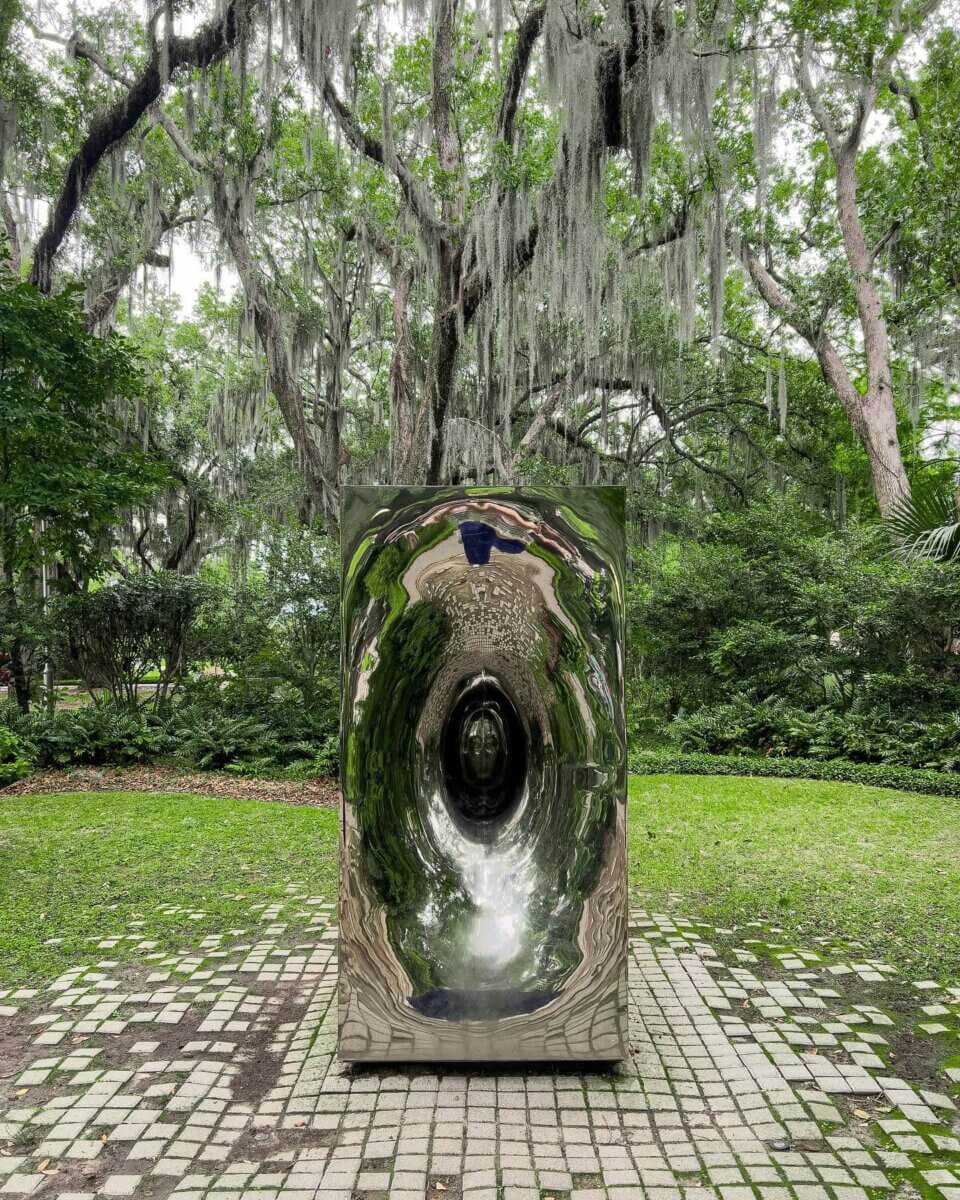 7 Southern Art Museums & Installations Worth Planning a Trip Around - 11 A tall, mirrored sculpture with a tunnel-like center stands on a stone path in a lush, green park with hanging moss at the New Orleans Museum of Art.