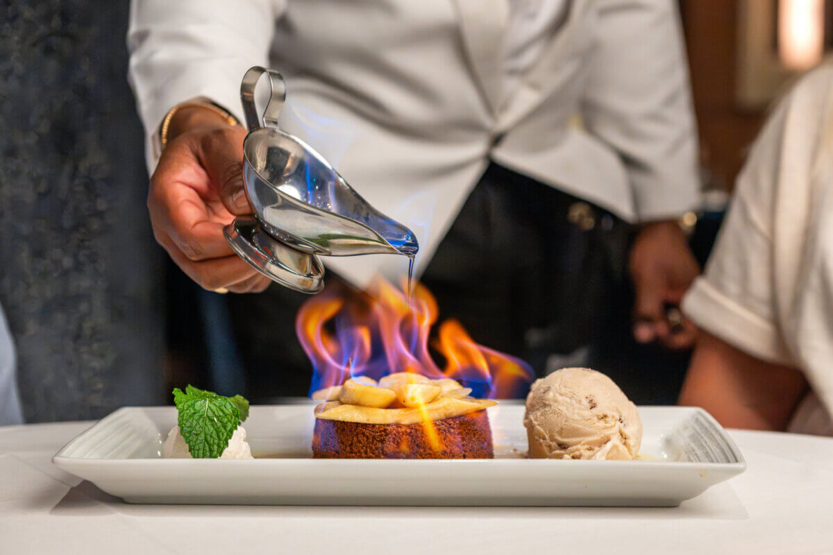 The Best Culinary Hot Spots in the South - 3 A server pours sauce over a dessert with bananas, which is on fire, next to a scoop of ice cream and a mint garnish on a white plate—an unforgettable experience at one of the South’s best culinary hot spots.
