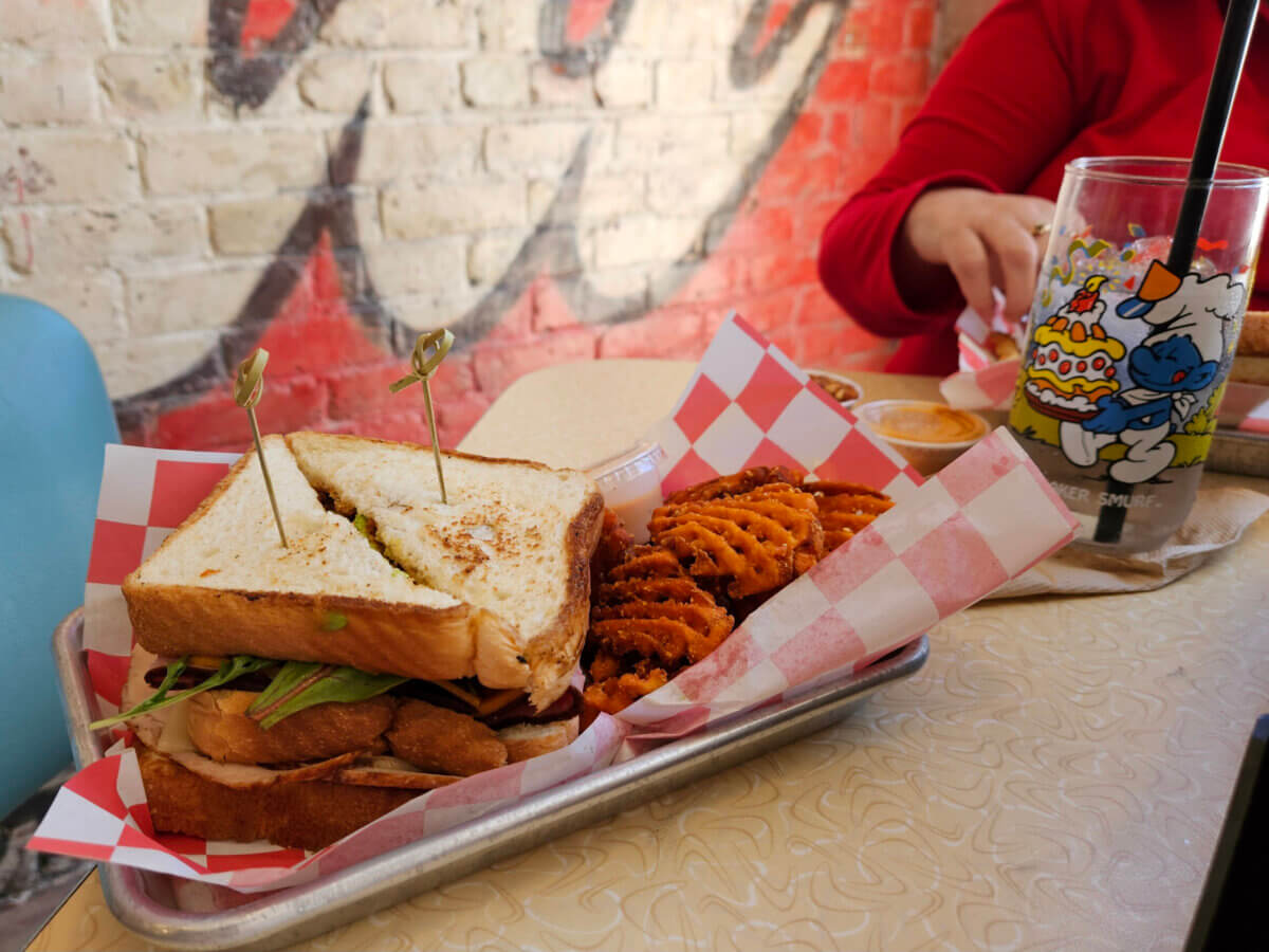 The Best Culinary Hot Spots in the South - 12 A tray with a sandwich cut in half and waffle fries on red-checked paper; a glass with a cartoon character and a person in a red shirt are in the background, showcasing the kind of classic comfort food you’d find at the best southern restaurants.