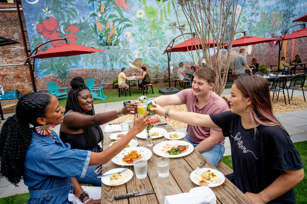The Best Culinary Hot Spots in the South - 4 Four people sit at an outdoor table, sharing a meal and toasting with drinks at one of the South’s best culinary hot spots. Other diners and a colorful mural are visible in the background.