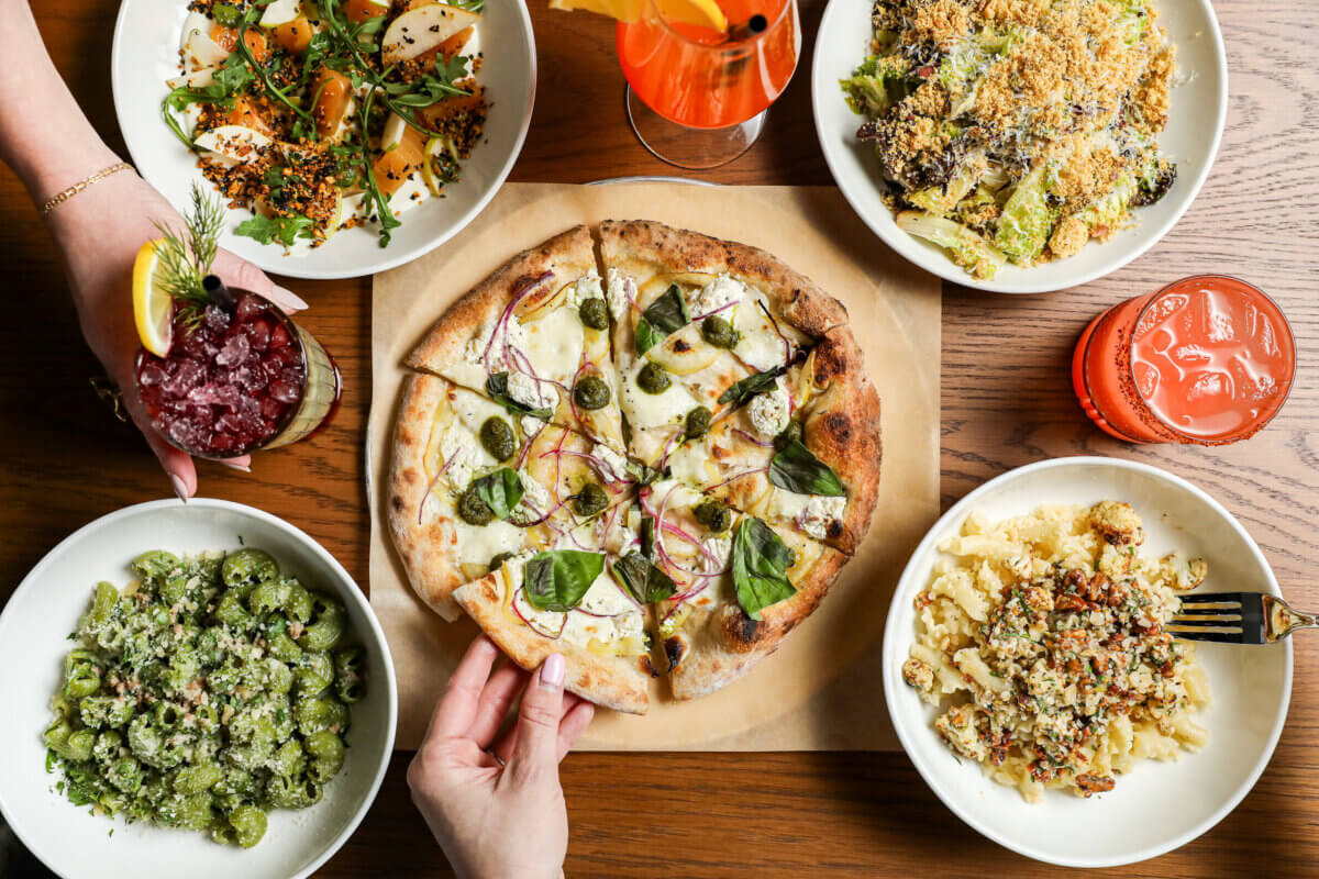 The Best Culinary Hot Spots in the South - 9 A table with a pizza, pasta dishes, salads, and two drinks, with hands reaching for food—capturing the lively atmosphere often found in culinary hot spots and some of the best restaurants.