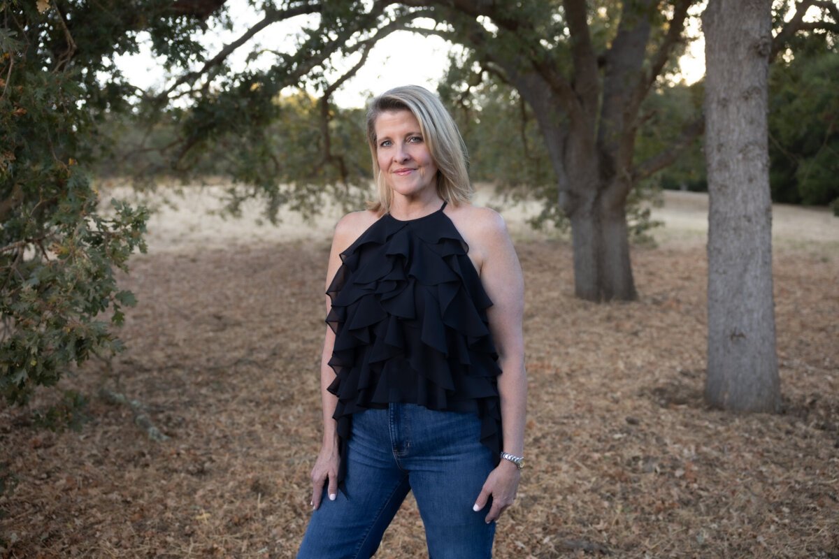 A woman stands outdoors on dry ground, wearing a sleeveless black ruffled top and blue jeans, with trees and grass in the background.