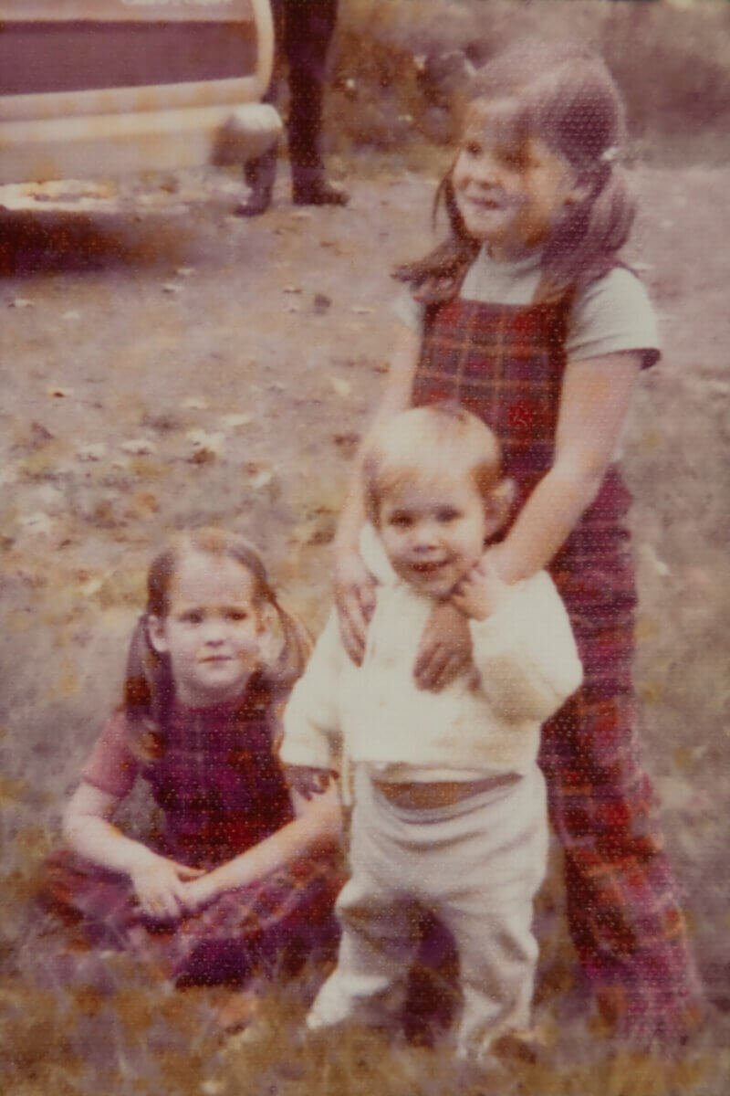 Three children pose outdoors; two girls in plaid outfits, one standing with hands on a toddler in a white sweater, while the other girl sits on the grass.