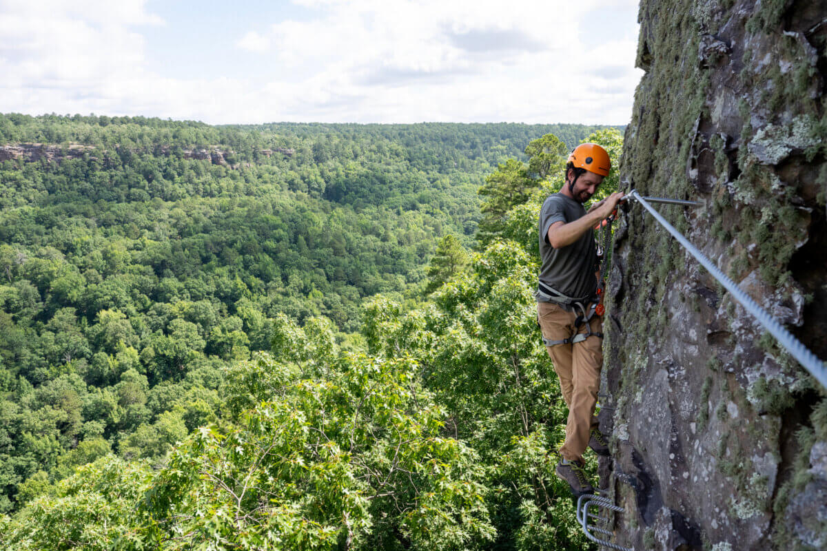 5 NEW Hikes to Explore Around the South - 2 Person wearing a helmet and harness climbs a rocky cliff face using safety cables, exploring the new via ferrata at Petit Jean State Park.