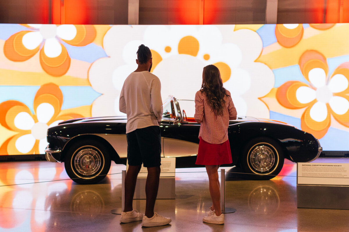 Two people stand in front of a black vintage car displayed indoors, with a bright, flower-patterned backdrop behind them—one of those southern attractions worth the drive.