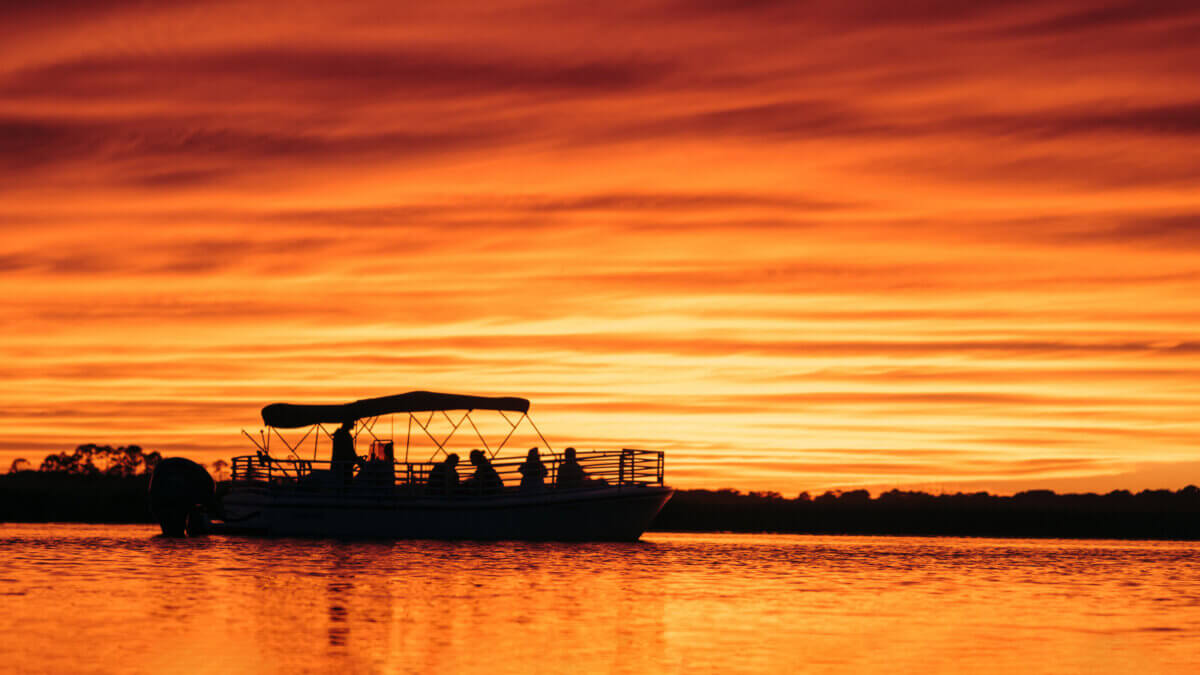Silhouetted boat with several people on calm water during a vibrant orange sunset, exuding Southern Charm as streaked clouds fill the sky.