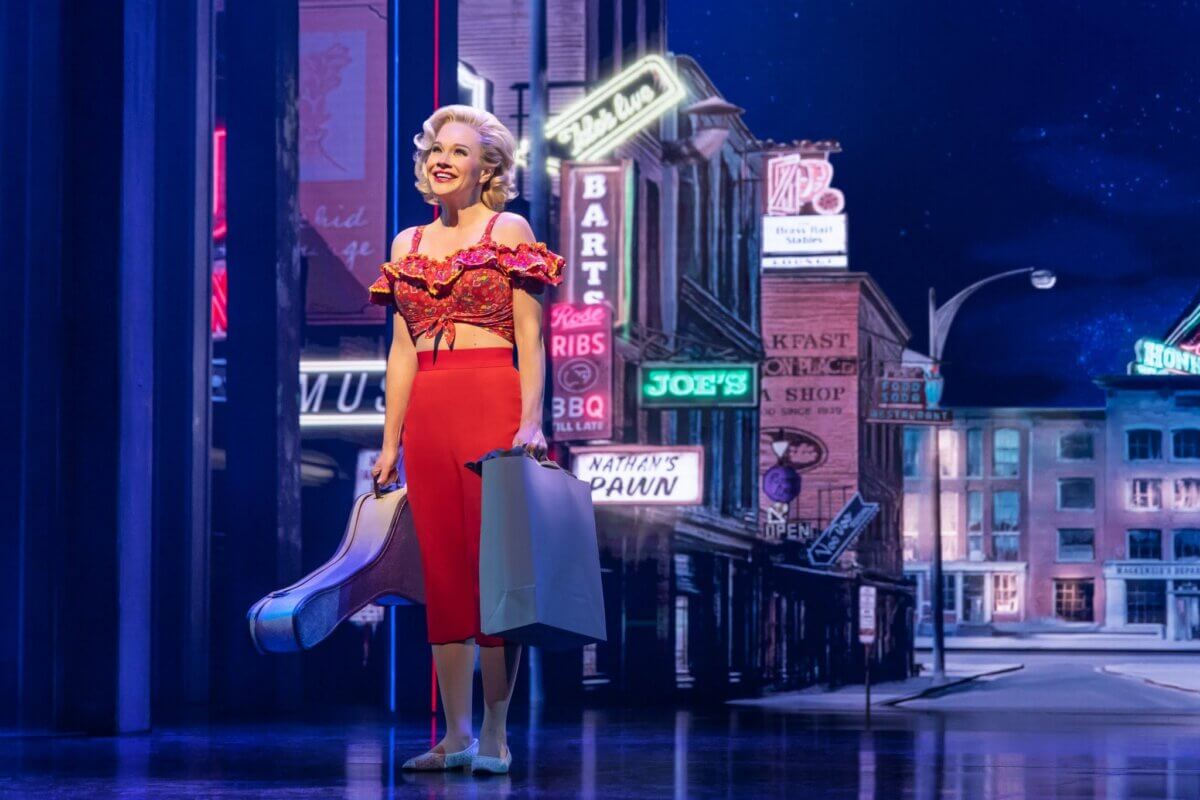 For the Love of Dolly: 7 Ways to Celebrate Her Legacy - 5 A woman in a red outfit stands on stage holding shopping bags and a guitar case, with a city street backdrop featuring neon signs behind her from "Dolly," a new musical headed to Broadway.