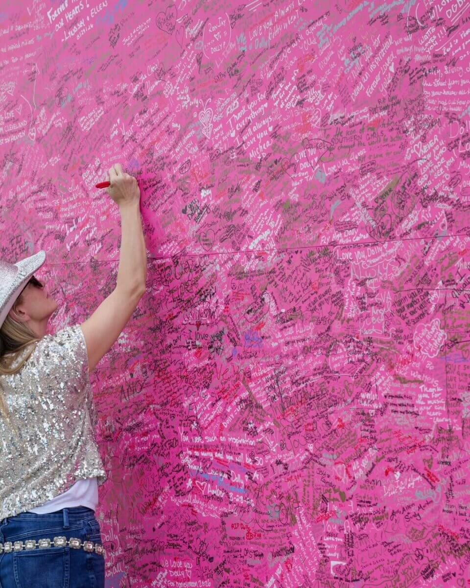 For the Love of Dolly: 7 Ways to Celebrate Her Legacy - 10 A person in a sequin top and hat writes on a large pink wall covered with handwritten messages in various colors, adding their own note to celebrate Dolly among the vibrant tributes.