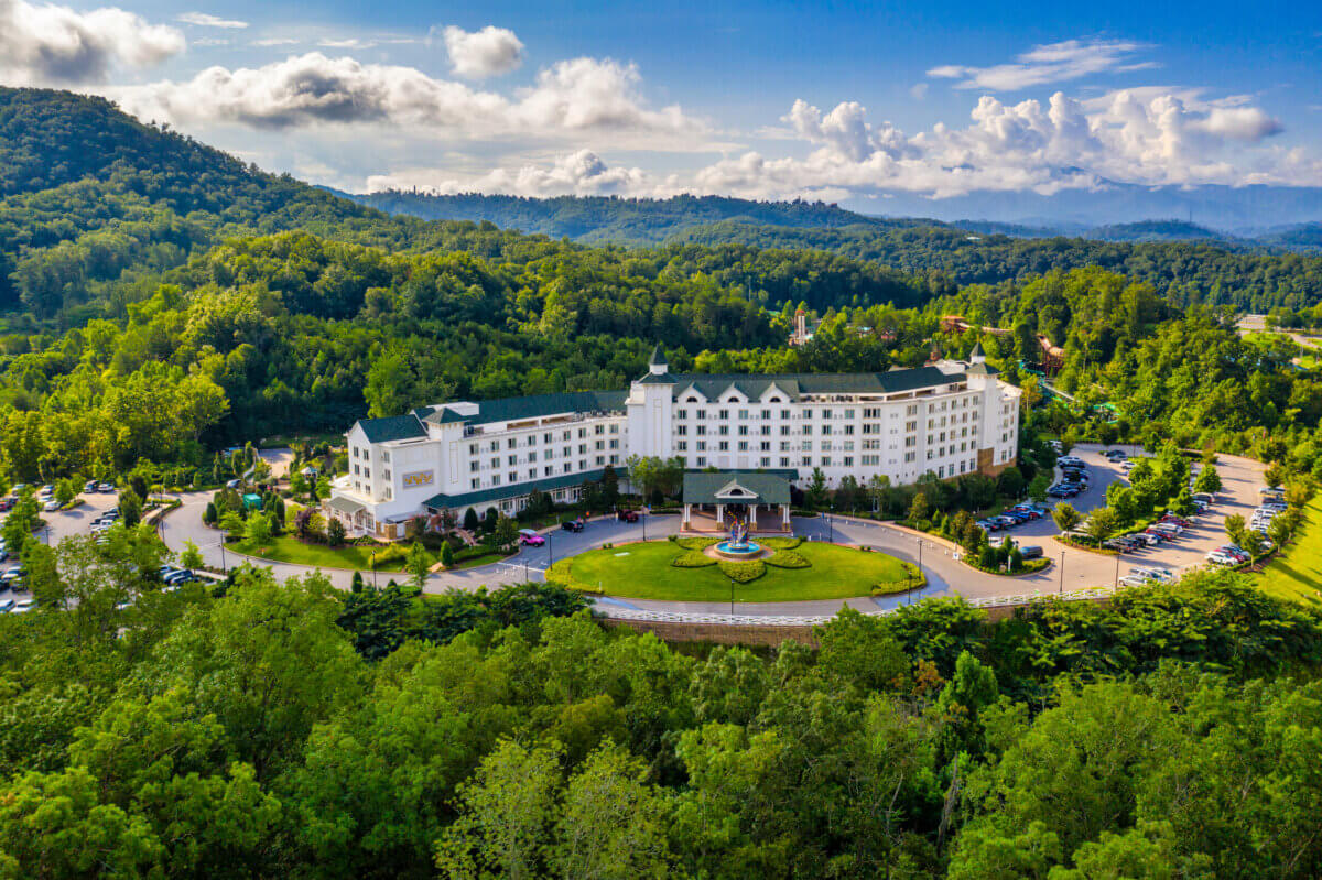 For the Love of Dolly: 7 Ways to Celebrate Her Legacy - 7 A large white hotel with a circular driveway and fountain, surrounded by trees and mountains under a partly cloudy sky, welcomes guests who are eager to celebrate Dolly in style or embark on unforgettable Dolly trips.