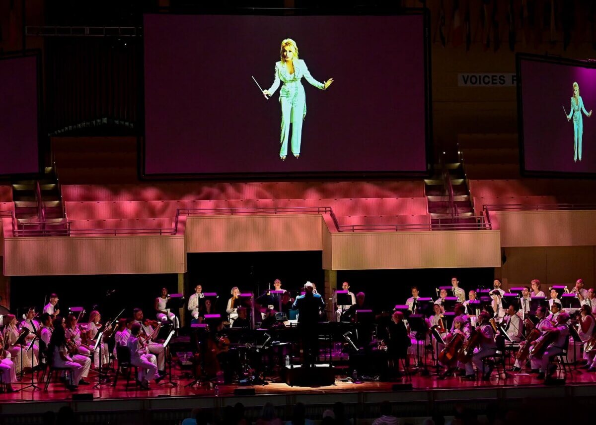 For the Love of Dolly: 7 Ways to Celebrate Her Legacy - 11 A symphony orchestra performs on stage with a large screen above showing Dolly Parton holding a conductor's baton at a production of Threads.