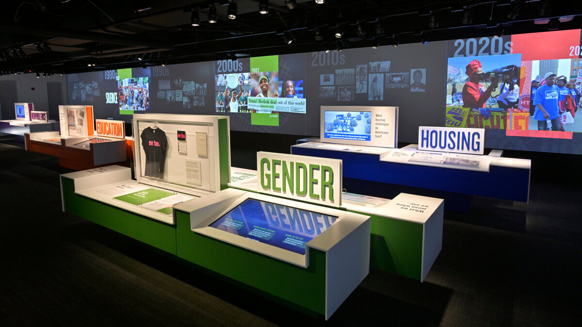 Museum exhibit with displays labeled “GENDER,” “EDUCATION,” and “HOUSING,” featuring text, artifacts, and large wall projections of historical images and dates—an immersive experience that stands out among Southern attractions and is truly worth the drive.