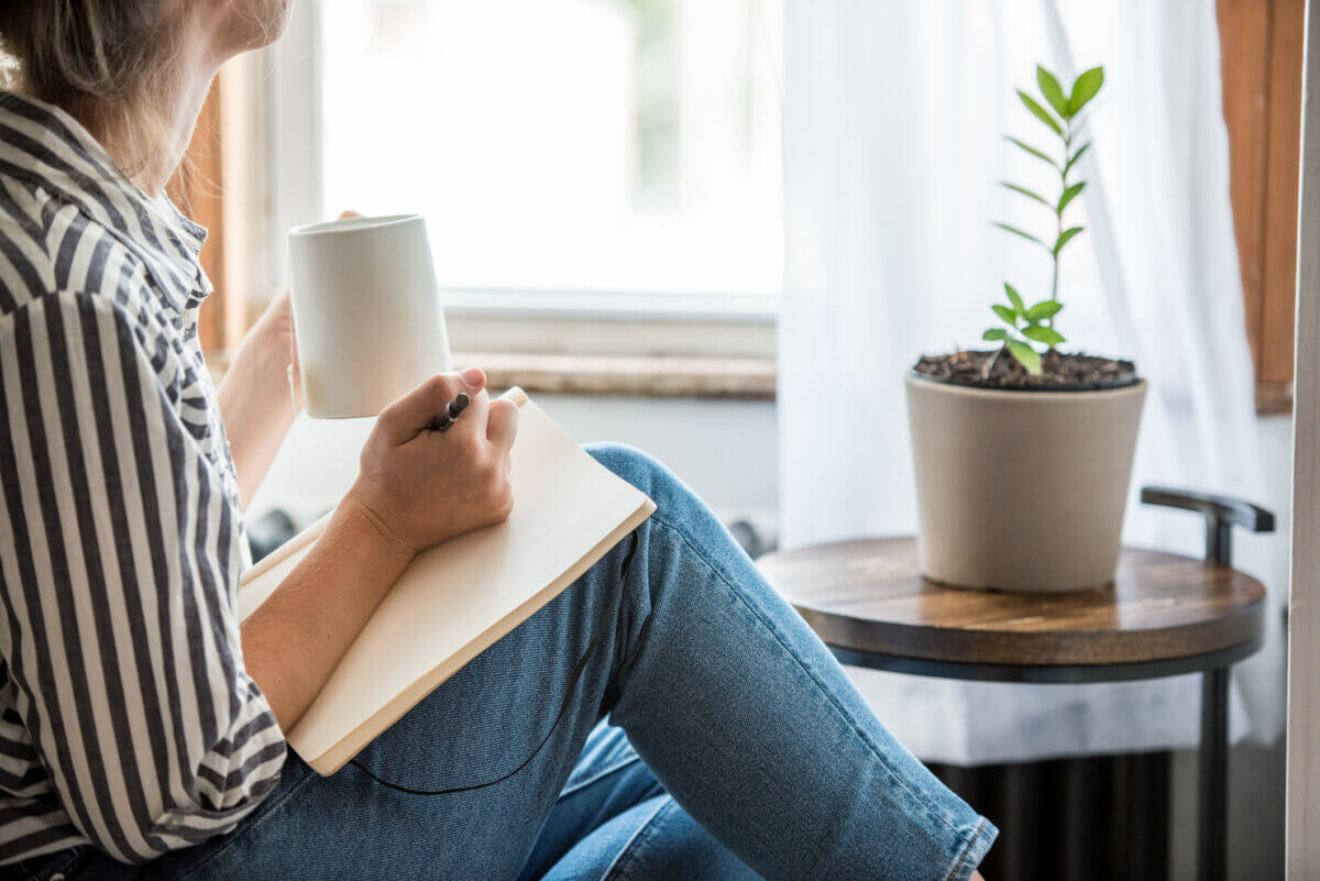 3 Tools to Move from Money Anxiety to Empowerment - 2 A person holding a mug and writing in a notebook sits by a window next to a potted plant on a small round table, thoughtfully reflecting on their goals with the help of money management tools.