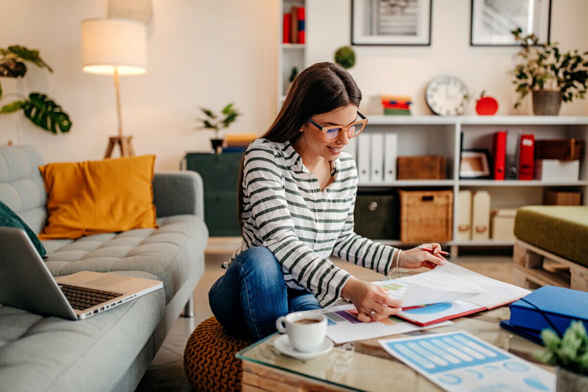 3 Tools to Move from Money Anxiety to Empowerment - 3 Woman sitting on the floor in a living room, working with documents and charts on a glass table, utilizing financial tools on her laptop to manage her money, with a cup of coffee nearby.