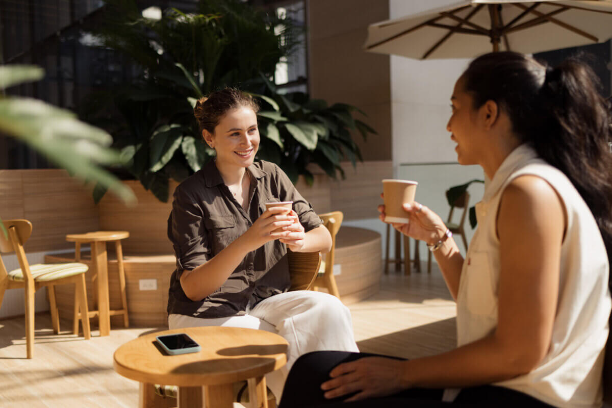 3 Tools to Move from Money Anxiety to Empowerment - 4 Two women sit at a small table in a bright, modern café, holding paper cups and talking. As they chat about ways to manage my money effectively, a smartphone—perhaps loaded with the latest money management tools—rests on the table between them.