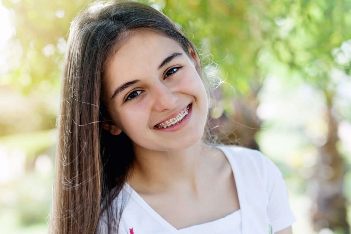 Healthy Smiles for Life: A Guide to Family Dental Care For Every Age - 5 Teen girl with long brown hair and braces smiles outdoors, wearing a white shirt, with blurred green foliage in the background—an image that embodies healthy smiles and highlights the positive results of family dental care.