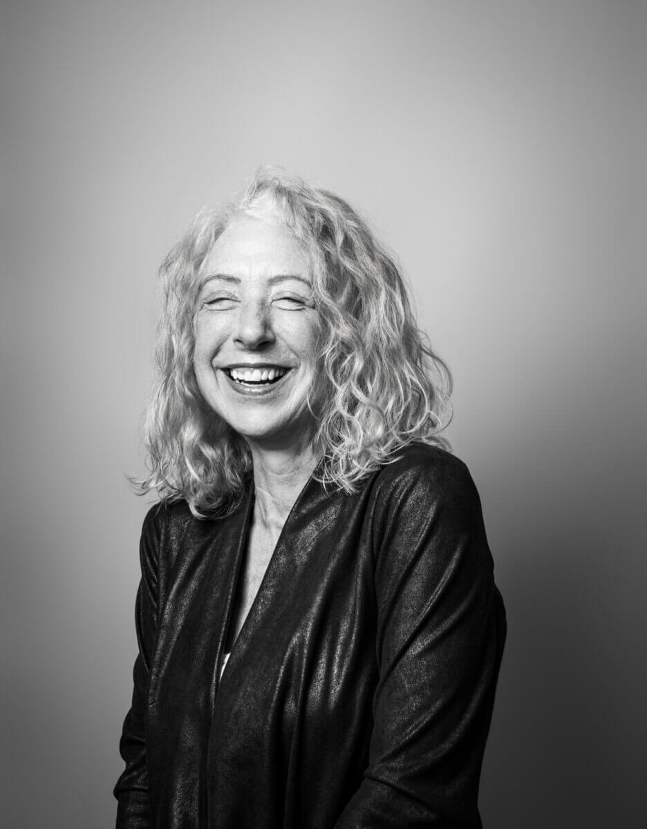 Black and white photo of a smiling older woman with curly hair wearing a dark jacket, standing against a plain light background. Inspired by her time volunteering with Nashville Cat Rescue, Molly Reed radiates warmth and compassion in this timeless portrait.