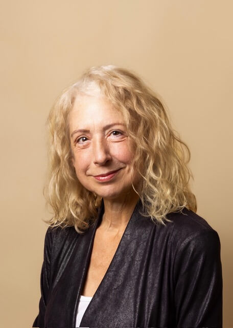 An older woman with curly blonde hair, wearing a dark jacket over a white top, stands against a plain beige background, looking at the camera and smiling slightly. Molly Reed, a supporter of Nashville Cat Rescue, exudes warmth and compassion in her gentle expression.