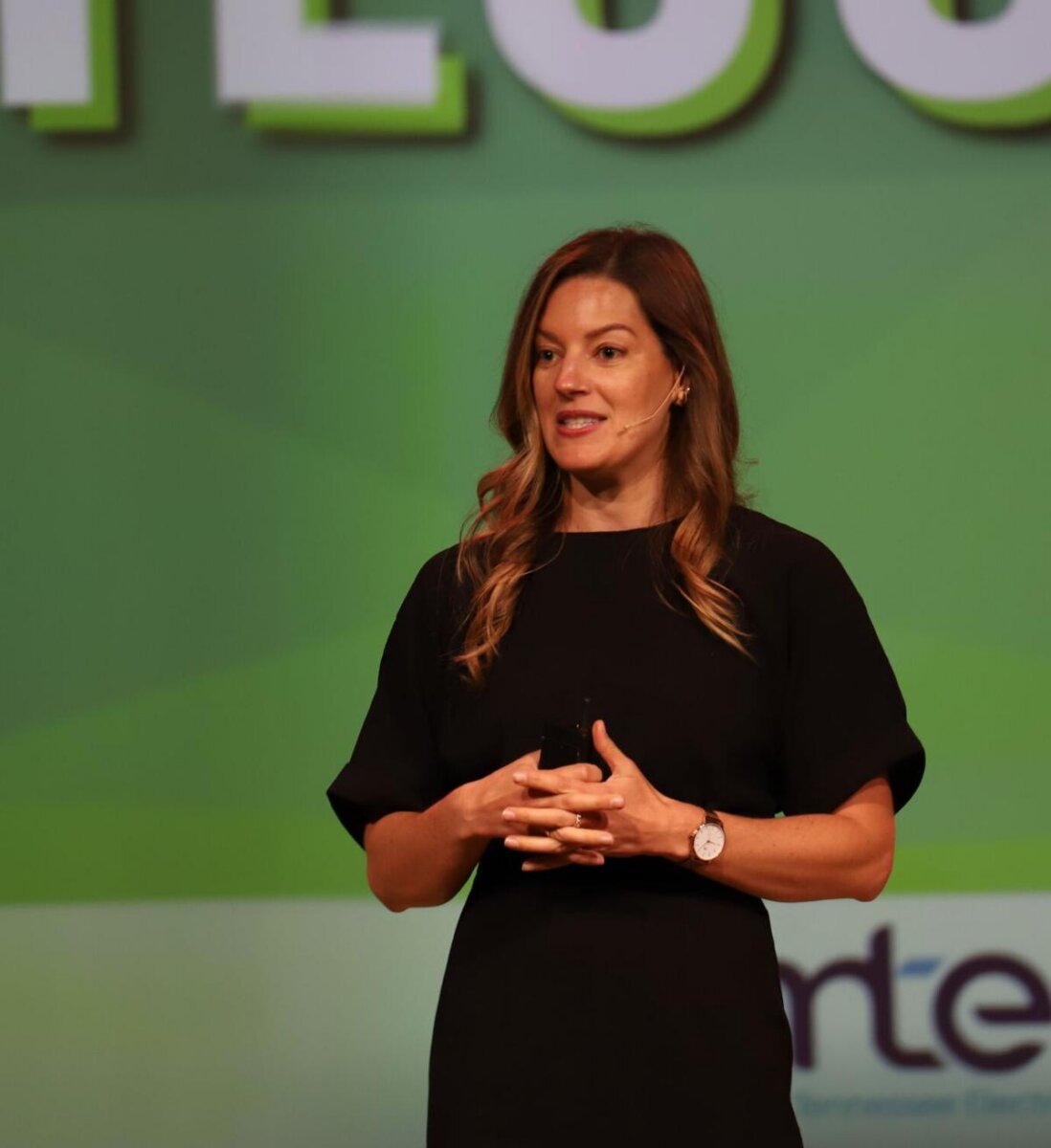 Laurel Graefe, a woman with long brown hair in a black dress, stands on stage speaking with hands clasped. A green and white background with large text is behind her.