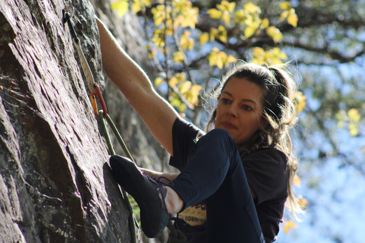 Woman climbing a rock face outdoors, secured with a harness and rope; sunlight filters through trees in the background.
