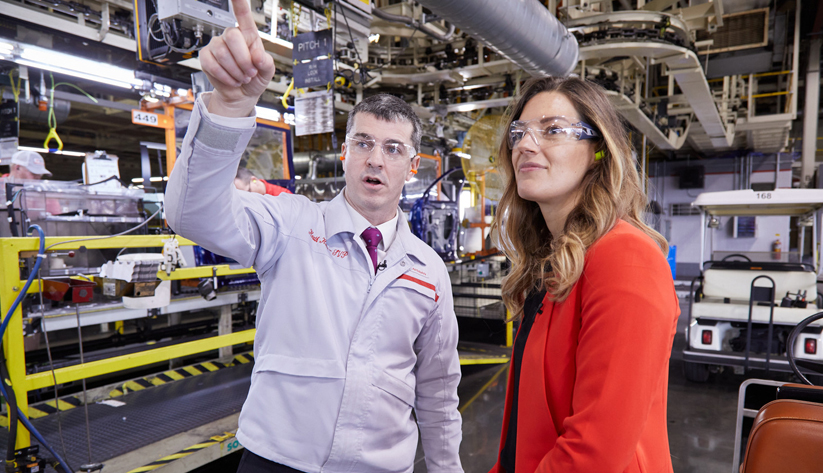 Two people wearing safety glasses and business attire stand in a factory, with one person pointing while explaining machinery in the background.