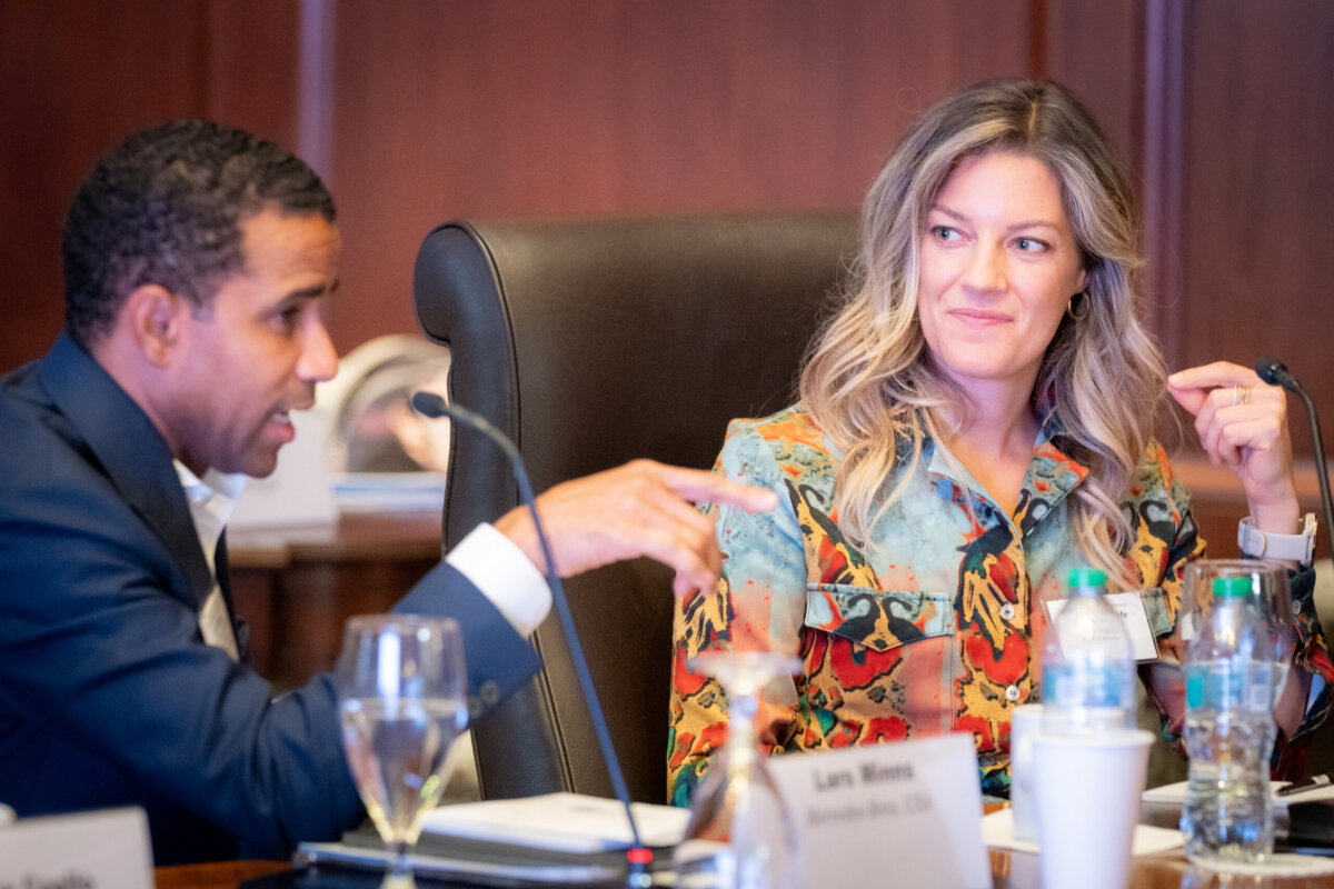 Two people sit at a conference table in discussion. One gestures while speaking, and the other listens attentively. Nameplates, water bottles, and papers are visible on the table.